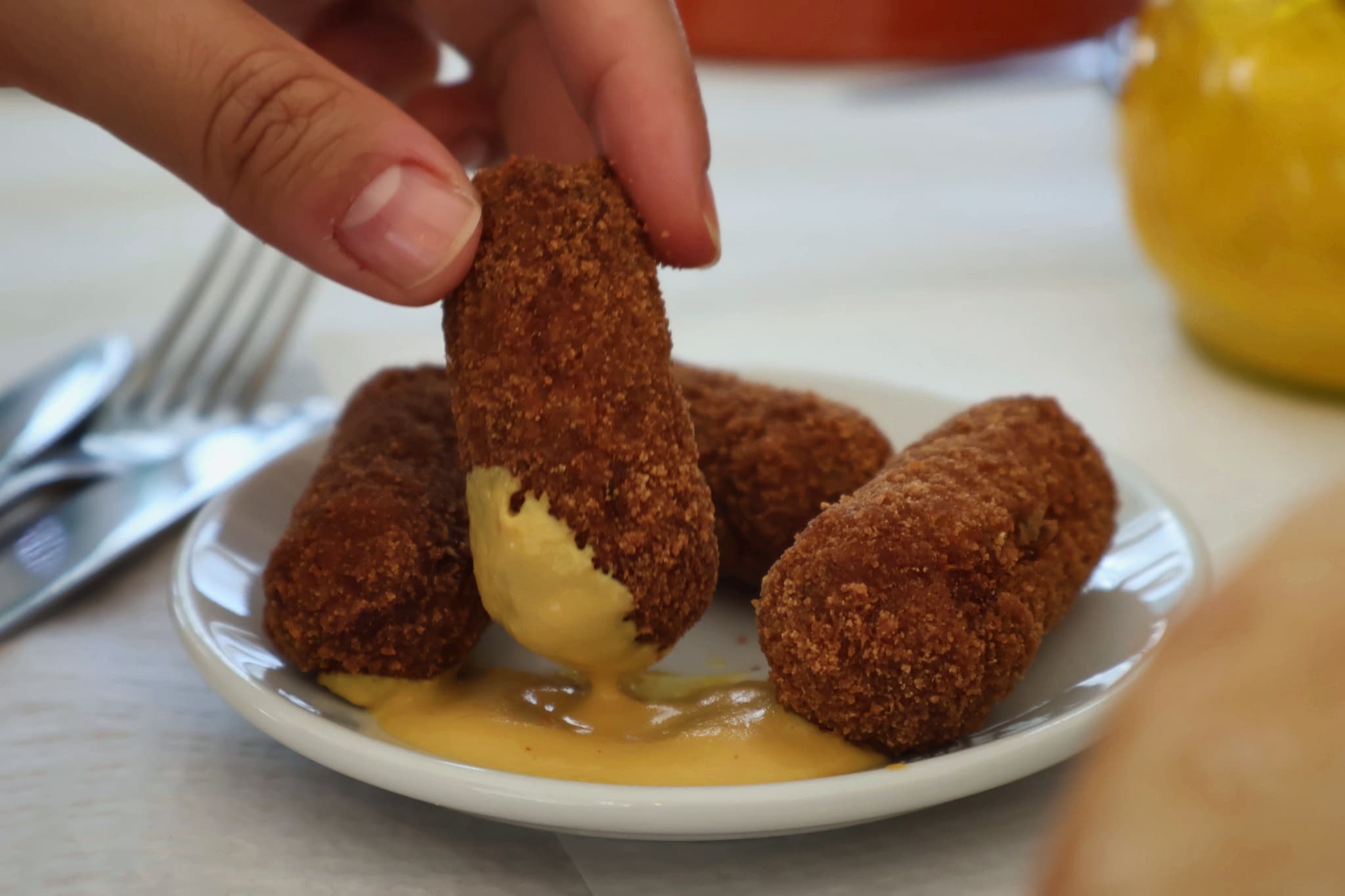 Mão a mergulhar um croquete em molho de mostarda, num prato branco.